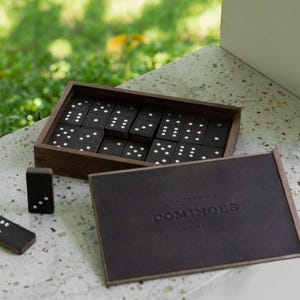 May include: A wooden domino set with black tiles and white dots, neatly arranged in a dark wooden box. The set includes a dark brown leather-bound box with the word "DOMINOES" embossed on the cover. The game is set on a light-colored surface.