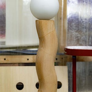 May include: A white sphere sits atop a wooden, curved pedestal. The pedestal is made of light brown wood with a smooth, polished finish.