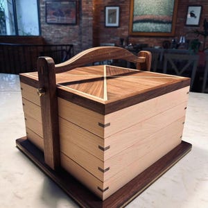 May include: A handcrafted wooden box with a handle, featuring a light wood body and dark wood accents. The lid has a geometric design, and the box is constructed with dovetail joints. The box sits on a rectangular base.