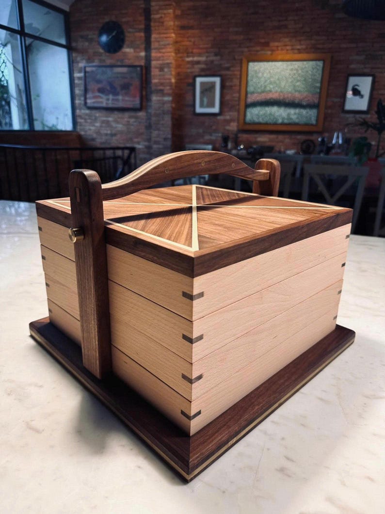 May include: A handcrafted wooden box with a handle, featuring a light wood body and dark wood accents. The lid has a geometric design, and the box is constructed with dovetail joints. The box sits on a rectangular base.