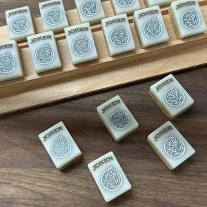 May include: Ivory-colored Mahjong tiles with the word "JOKER" and a decorative emblem. The tiles are arranged on a wooden surface, some in a wooden tray. The tiles are rectangular and feature a detailed design.