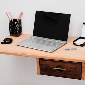 May include: A light wood floating desk with a drawer. The drawer is made of dark wood with a brass handle. A laptop, a pencil holder, a notepad, and a pen are on the desk.