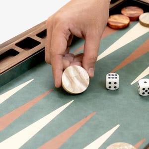 May include: A backgammon board with a green playing surface, brown and white triangles, and wooden game pieces. Two white dice with black dots are visible. A hand is placing a light-colored wooden game piece on the board.