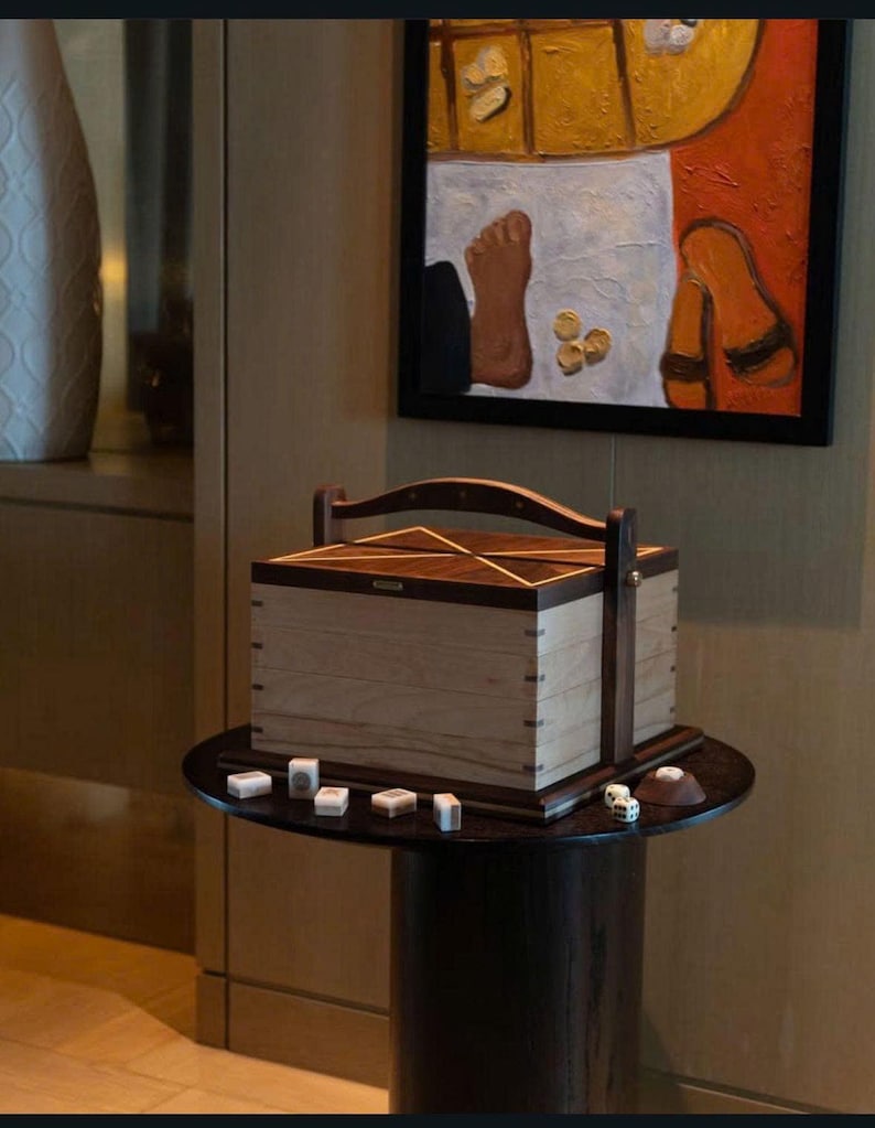 May include: A wooden box with a handle and a geometric design sits on a small, dark round table. Dice and game tiles are scattered around the box. A painting with a foot and sandals hangs in the background.