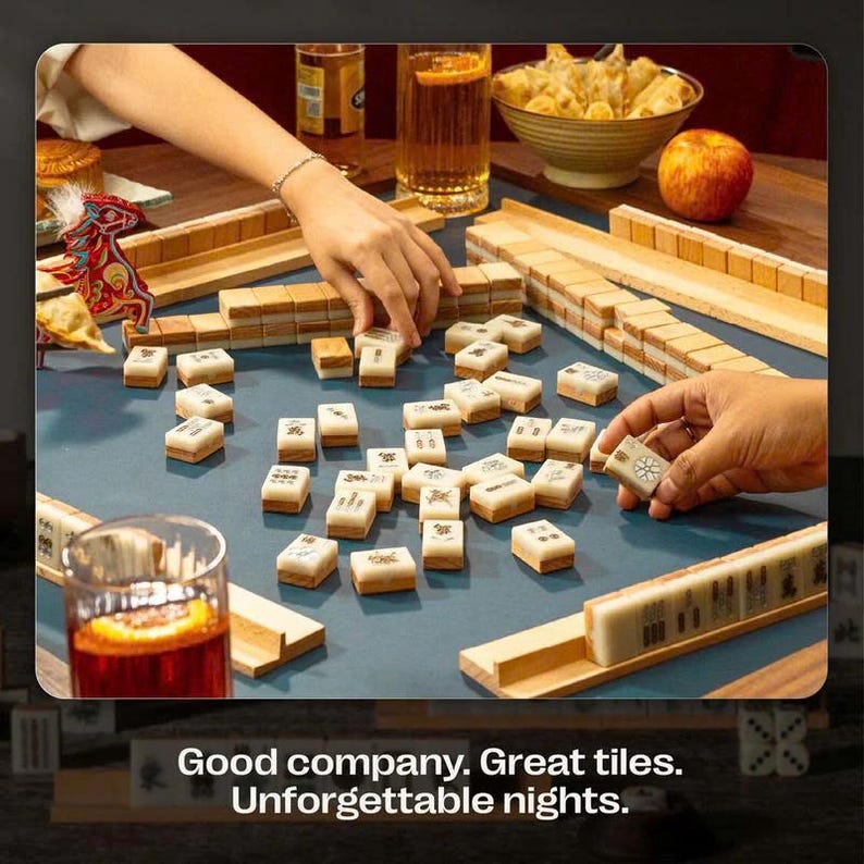 May include: A game of Mahjong in progress, with hands reaching for tiles on a blue surface. The tiles are beige with black symbols. The scene includes drinks, snacks, and the text "Good company. Great tiles. Unforgettable nights."