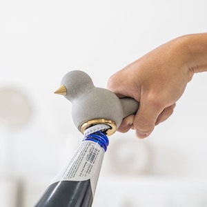 Puede incluir: Un abridor de botellas de pájaro de hormigón gris con una parte superior de metal dorado se está utilizando para abrir una botella de cerveza.