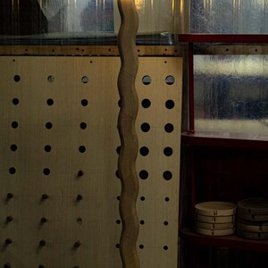 May include: A wooden floor lamp with a white globe light. The lamp has a wavy, organic shape and a black base. The lamp is in a workshop setting with a pegboard wall and a shelf in the background.