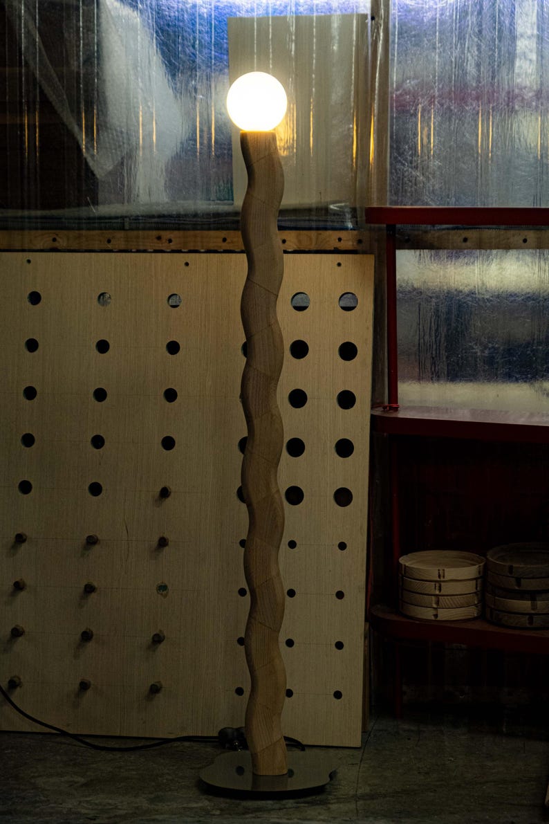 May include: A wooden floor lamp with a white globe light. The lamp has a wavy, organic shape and a black base. The lamp is in a workshop setting with a pegboard wall and a shelf in the background.