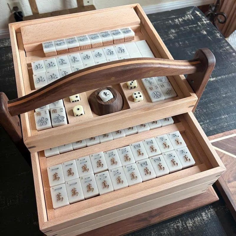 May include: A wooden Mahjong set with a two-tiered design. The top tier holds tiles, dice, and a wooden tile holder. The bottom tier is filled with more tiles. The set has a dark wooden handle and is displayed on a dark wooden surface.