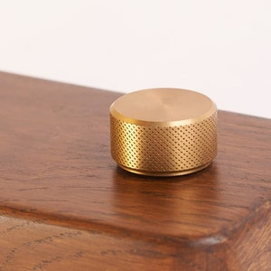 May include: A gold-coloured knob with a textured surface. The knob is sitting on a wooden surface.