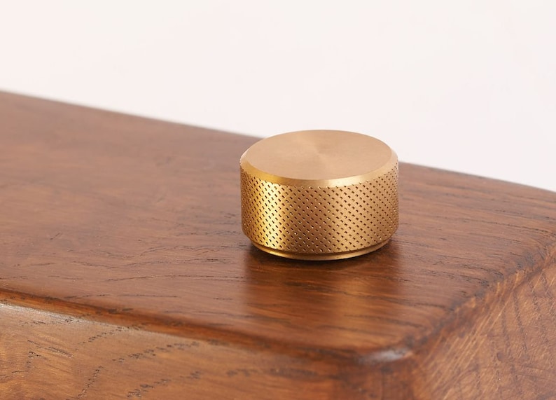 May include: A gold-coloured knob with a textured surface. The knob is sitting on a wooden surface.