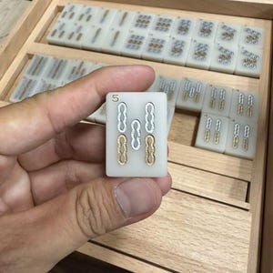 May include: A close-up of a Mahjong tile held in a hand, displaying the number "5" at the top. The tile features a white background with gold and silver markings. Other tiles are visible in a wooden tray.