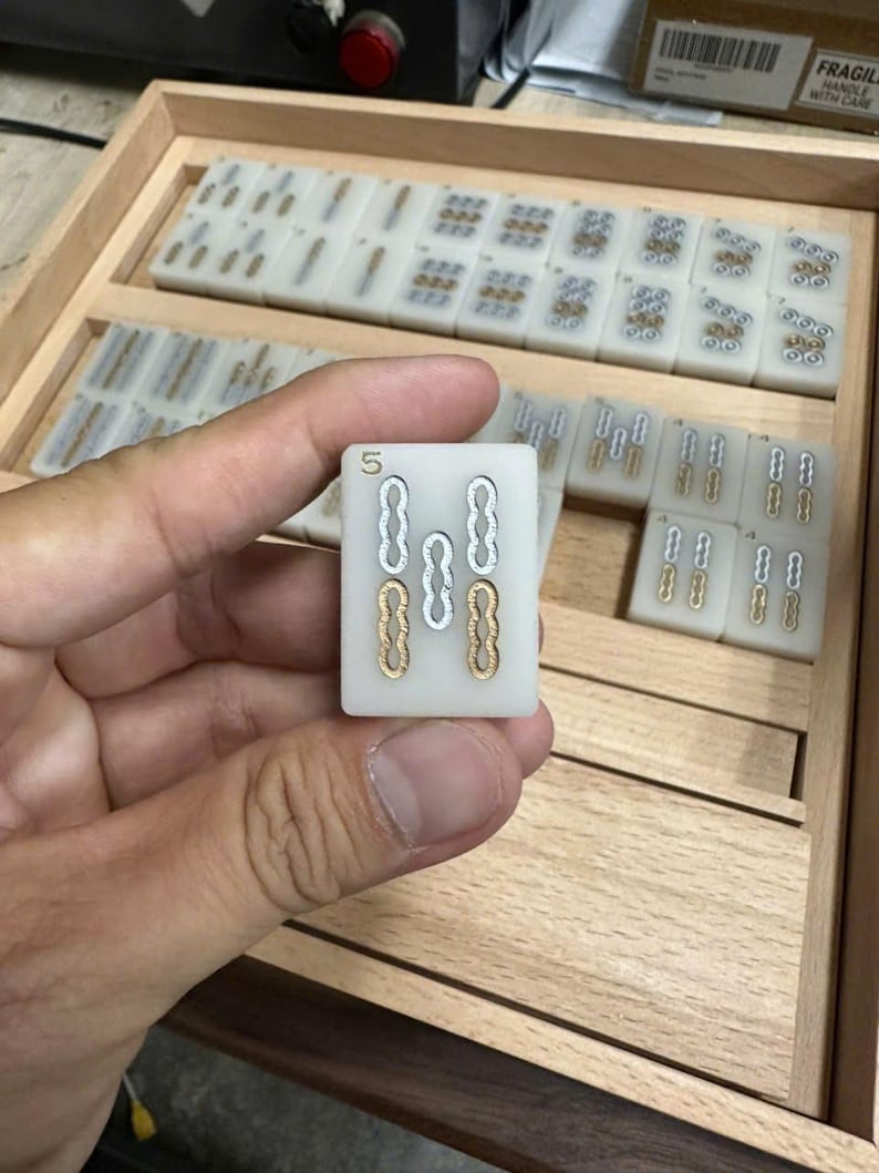 May include: A close-up of a Mahjong tile held in a hand, displaying the number "5" at the top. The tile features a white background with gold and silver markings. Other tiles are visible in a wooden tray.