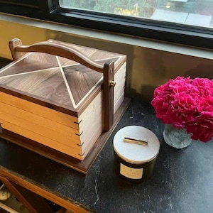 May include: A wooden box with a handle, featuring a geometric design on the lid. The box is made of light and dark wood, with a handle for carrying. A candle in a glass jar and a vase of pink flowers are also visible.