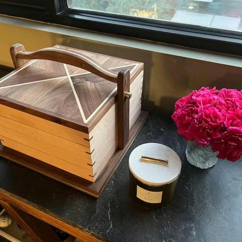 May include: A wooden box with a handle, featuring a geometric design on the lid. The box is made of light and dark wood, with a handle for carrying. A candle in a glass jar and a vase of pink flowers are also visible.