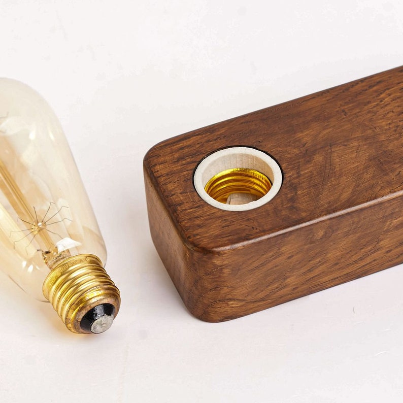 May include: A wooden light fitting with a single light bulb socket. The socket is made of white ceramic and is surrounded by a dark brown wooden frame. The light bulb is a vintage-style filament bulb.