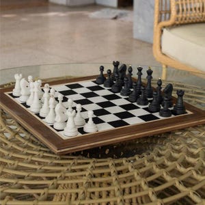 May include: A wooden chess set with black and white pieces arranged on a checkered board. The board is set in a wooden frame. The chess pieces are black and white. The set is on a woven table.