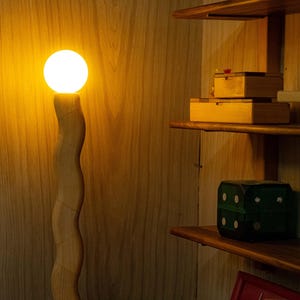 May include: A wooden floor lamp with a white light bulb. The lamp is in a room with wooden shelves and a green box on the shelf.