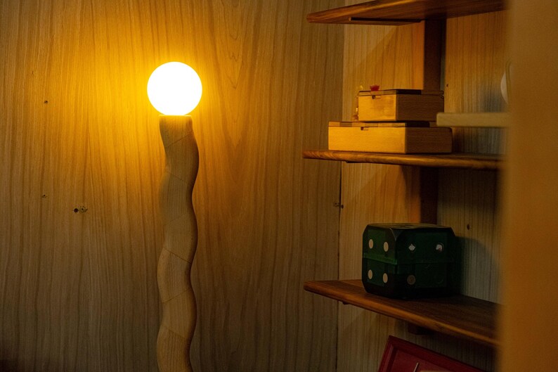 May include: A wooden floor lamp with a white light bulb. The lamp is in a room with wooden shelves and a green box on the shelf.