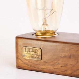May include: A wooden lamp base with a brass light socket and a clear glass light bulb. The base has a brass plaque that reads "LUXURY crafted in Virginia 0202/1000".