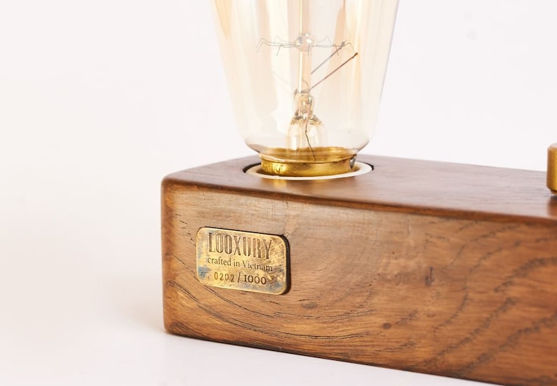 May include: A wooden lamp base with a brass light socket and a clear glass light bulb. The base has a brass plaque that reads "LUXURY crafted in Virginia 0202/1000".