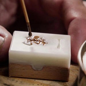 May include: Close-up of a jeweler working on a small gold-colored object. The object is in a white and brown mold, with a brush applying a liquid. A gold-colored bowl is in the background.