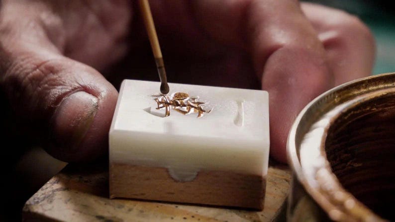 May include: Close-up of a jeweler working on a small gold-colored object. The object is in a white and brown mold, with a brush applying a liquid. A gold-colored bowl is in the background.