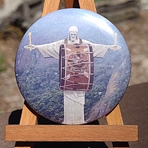May include: A round button featuring a white statue of a person with outstretched arms holding a drum. The statue is set against a blue sky and green mountains. The drum has a red and brown design with the text "The Essential Band" printed on it.