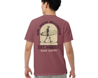 May include: A maroon t-shirt with a graphic of a skeleton surfing on a wave. The graphic is surrounded by a circle with the text "Surf Break Beachwear Est. MMXXIII Bone Surfer".