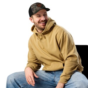 May include: A person wearing a tan hoodie and blue jeans. They are wearing a black and green camouflage trucker hat with a white and orange logo that says "Never Give Up".