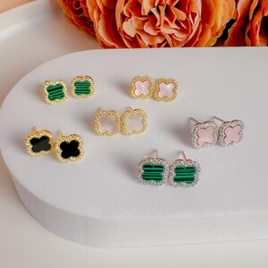 May include: A collection of clover-shaped stud earrings in various colors, including green, pink, black, and white. The earrings have gold or silver-colored frames and are displayed on a white surface. Some earrings are adorned with small, clear crystals.
