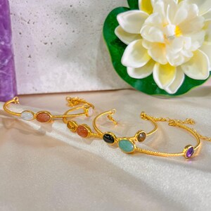Könnte beinhalten: Goldfarbene Armbänder mit bunten Edelsteinen. Jedes Armband hat eine zarte Kette und ist mit oval geformten Steinen in verschiedenen Farben wie Weiß, Orange, Schwarz und Lila verziert. Die Armbänder werden auf einer satinartigen Oberfläche präsentiert.