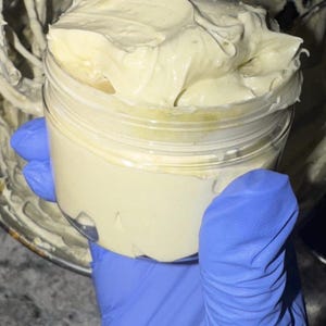 May include: A clear plastic jar filled with a creamy, off-white substance. The jar is held by a person wearing blue gloves. The cream has a thick, whipped texture, and the jar is partially filled.