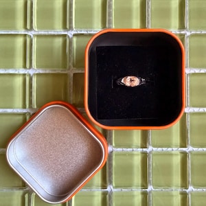 May include: A silver-toned ring featuring a central oval-shaped peach-colored gemstone, presented inside a square orange metal container with a black velvet insert and a separate matching orange lid with a metallic interior finish.