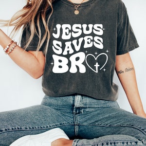 May include: A dark gray t-shirt with the text "JESUS SAVES BRO" in white with a heart and cross graphic.