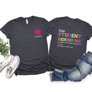 May include: Two dark gray t-shirts with a teacher-themed design. The front shirt has a pink heart with the text "My Student". The back shirt has the text "Dear Student Behind Me. The world is a better place with you in it. Love, The teacher in front of you." in rainbow colors.