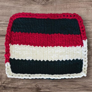 May include: A handmade, rectangular, knitted blanket with a striped pattern. The blanket features alternating bands of dark green, red, and white. The edges are trimmed with a thick red border, creating a cozy and textured appearance. The blanket is designed for comfort and warmth.