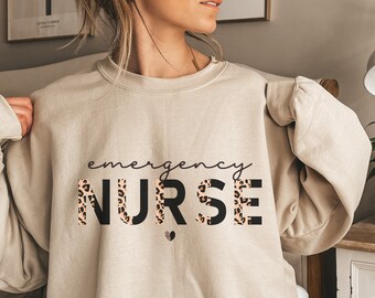 May include: A beige sweatshirt with the text "emergency NURSE" in black letters with a leopard print pattern.