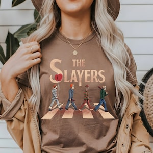 Op de afbeelding: Een bruin t-shirt met een afbeelding van vier horrorfilmfiguren die over een zebrapad lopen. De tekst "The Slayers" staat boven de figuren.