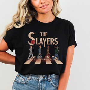 Op de afbeelding: Zwart T-shirt met een afbeelding van horrorfilmfiguren die over een zebrapad lopen. De tekst "The Slayers" staat boven de figuren.