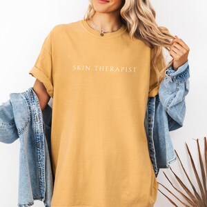 K&ouml;nnte beinhalten: Ein senfgelbes T-Shirt mit dem Aufdruck "SKIN THERAPIST" in Wei&szlig; auf der Vorderseite.