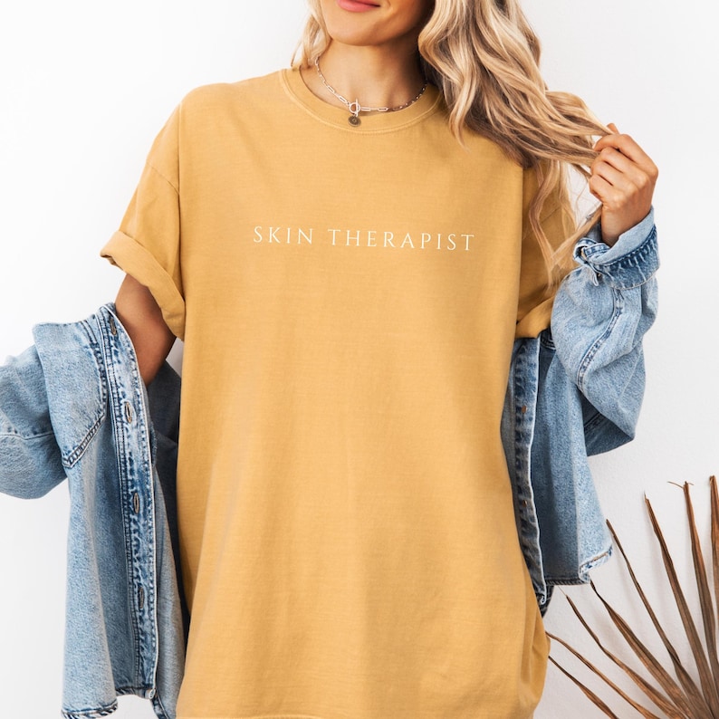 K&ouml;nnte beinhalten: Ein senfgelbes T-Shirt mit dem Aufdruck "SKIN THERAPIST" in Wei&szlig; auf der Vorderseite.
