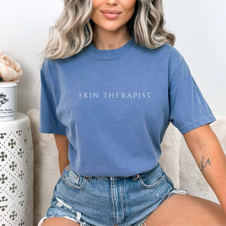 K&ouml;nnte beinhalten: Eine Frau tr&auml;gt ein blaues T-Shirt mit dem Aufdruck "SKIN THERAPIST". Sie tr&auml;gt Jeans-Shorts und hat ihre Haare offen.