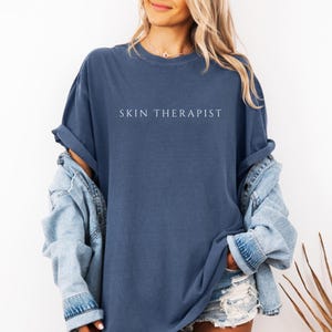 K&ouml;nnte beinhalten: Eine Frau tr&auml;gt ein blaues, &uuml;bergro&szlig;es T-Shirt mit dem Aufdruck "SKIN THERAPIST" in wei&szlig;. Sie tr&auml;gt eine Jeansjacke und eine Jeans-Shorts.