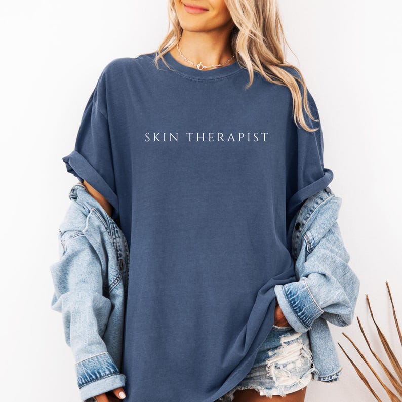 K&ouml;nnte beinhalten: Eine Frau tr&auml;gt ein blaues, &uuml;bergro&szlig;es T-Shirt mit dem Aufdruck "SKIN THERAPIST" in wei&szlig;. Sie tr&auml;gt eine Jeansjacke und eine Jeans-Shorts.