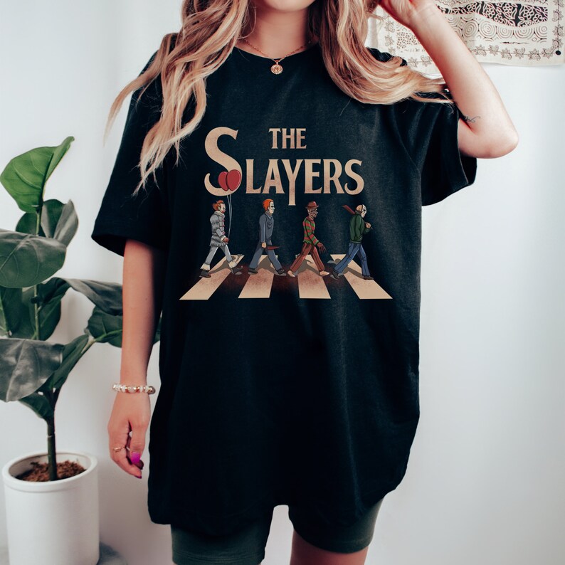 Op de afbeelding: Zwart T-shirt met een afbeelding van horrorfilmfiguren die over een zebrapad lopen. De tekst "The Slayers" staat boven de figuren.