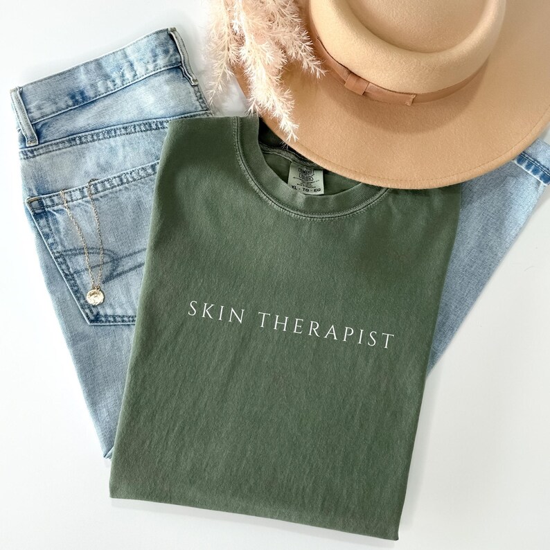 K&ouml;nnte beinhalten: Ein gr&uuml;nes T-Shirt mit dem Text "SKIN THERAPIST" in wei&szlig;en Buchstaben. Das T-Shirt liegt auf einem hellblauen Jeansstoff.