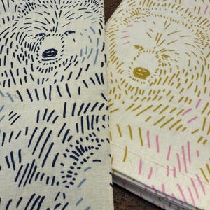 May include: Two folded linen napkins with bear illustrations. One napkin features a navy blue and light blue bear design on an off-white background. The other has a gold and pink bear design on an off-white background.