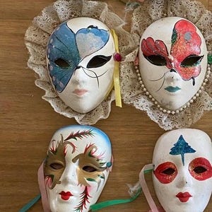 May include: A collection of decorative Venetian masks. Each mask is white with painted details, including red lips, eye designs, and colorful accents. Some masks have lace or ribbon trim.