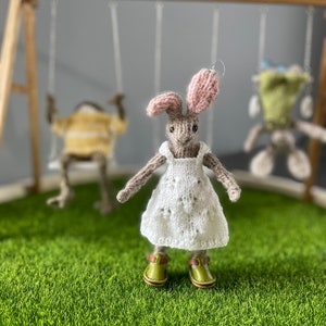 May include: A knitted bunny wearing a white dress with a floral pattern and green shoes. The bunny is standing on a green surface with a wooden swing set in the background.